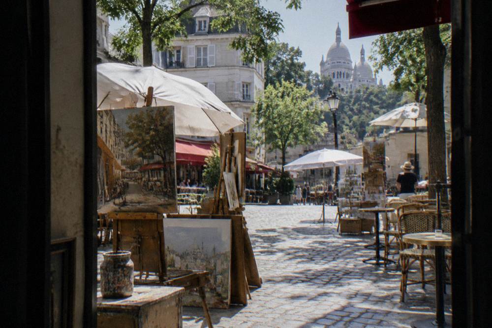 A city square with easels and painted canvases set out beside cafe tables
