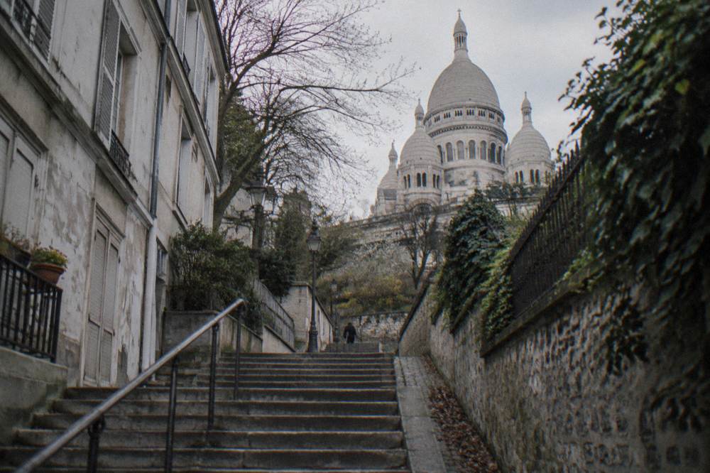 A stone staircase between old buildings climbing a Paris hill toward a domed basilica