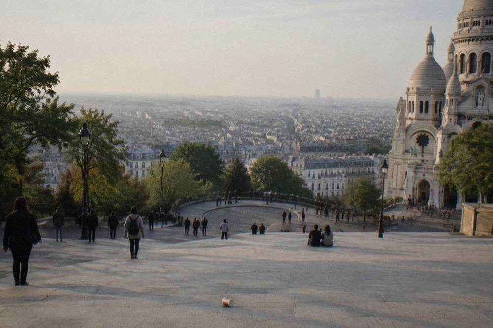 Wide stone forecourt in front of the Sacre-Coeur basilica under a partly cloudy sky, visitors scattered across the steps