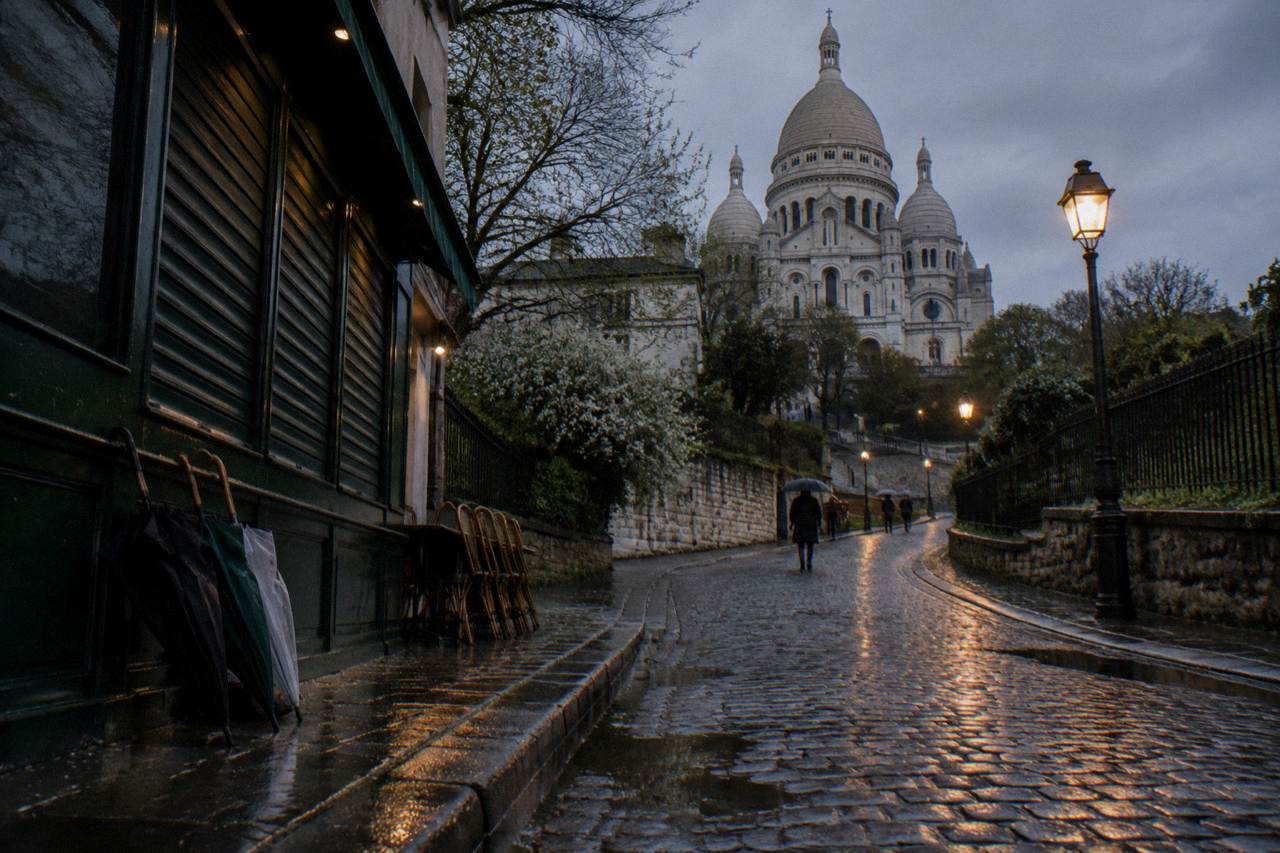 Rain-slicked cobblestone street in Montmartre curving uphill toward the basilica, umbrellas leaning against a shuttered cafe front