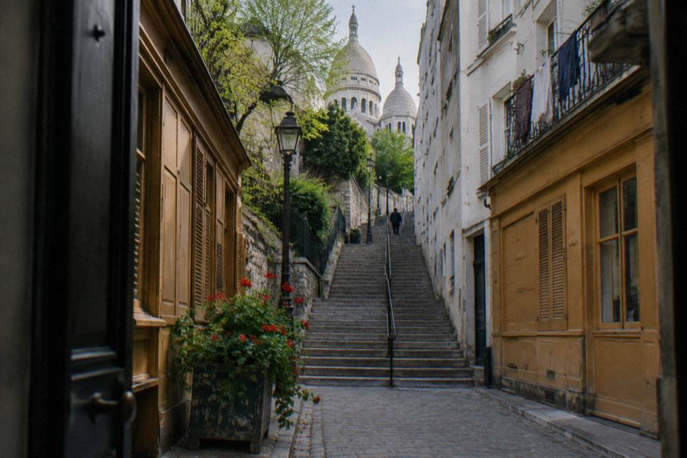 Narrow Montmartre street with stone steps climbing past shuttered shopfronts toward daylight