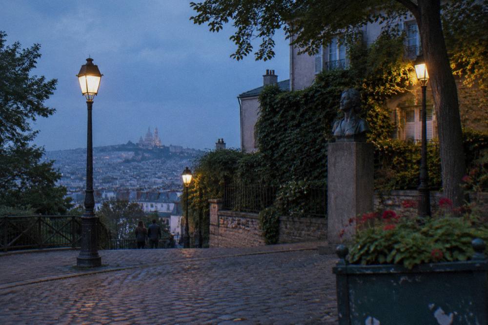 Street lamps glowing along a cobbled Montmartre lane at dusk with the city lit up below