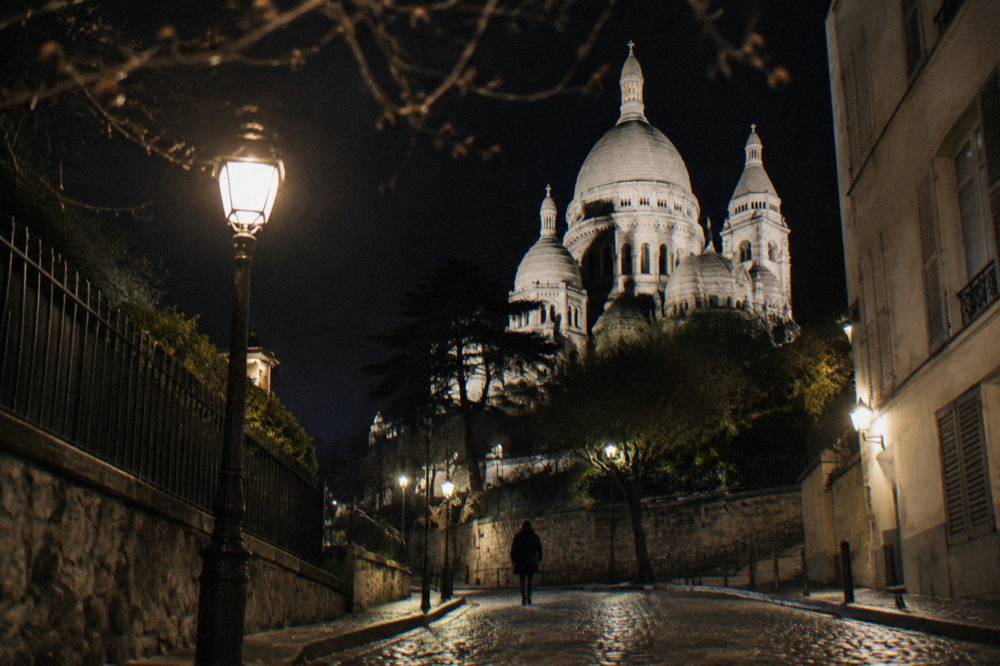 The illuminated white dome of Sacre-Coeur seen from a lamplit Montmartre street at night