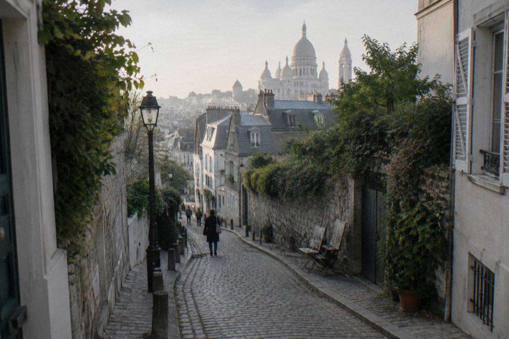 Narrow cobbled Montmartre street curving downhill with the basilica dome rising behind the rooftops